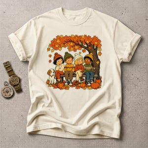 Puede incluir: Una camiseta de color crema con una ilustración gráfica de cuatro niños y un perro pequeño sentados en un banco bajo un árbol de otoño con hojas naranjas. Hay calabazas esparcidas por el suelo. Un reloj de pulsera está a la izquierda.