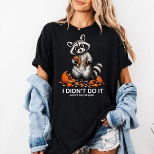 Puede incluir: Una camiseta negra con un gráfico de un mapache sosteniendo un trozo de calabaza naranja, rodeado de trozos de calabaza. El texto dice I DIDN'T DO IT y ...and I'll deny it again en letras mayúsculas blancas debajo de la ilustración.