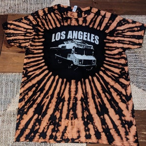 May include: Tie-dye t-shirt with a black and orange spiral pattern. The shirt features a black circle with the words "LOS ANGELES" above a graphic of a taco truck. The shirt is made of a soft material.