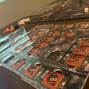 May include: A clear acrylic game organizer with brown and black card slots and a black and white game board with the text "By Day, Red By Night".