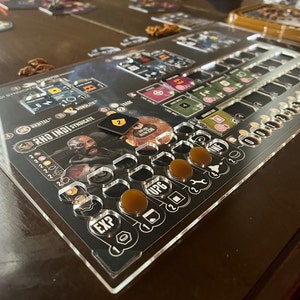 May include: A clear acrylic player board for the board game "Rho Indi Syndicate". The board features a character card, a shield token, and various spaces for tokens and dice. The board is labelled with the game's title and has spaces for experience points, upgrades, and other game elements.