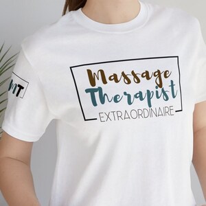 Op de afbeelding: Wit T-shirt met zwarte tekst die "Massage Therapist EXTRAORDINAIRE" in een kader zegt.