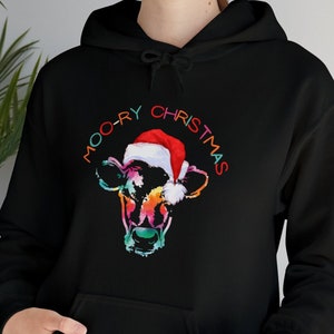 May include: Black hoodie featuring a colorful cow face wearing a Santa hat. The text "MOO-RY CHRISTMAS" arches above the cow's head in rainbow colors. The hoodie has a front pocket.