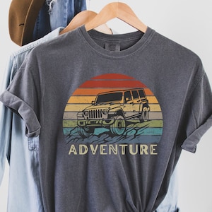 Może przedstawiać: Szary t-shirt z grafiką w stylu vintage przedstawiającą Jeepa jadącego przez krajobraz zachodu słońca. Pod grafiką znajduje się napis "ADVENTURE".
