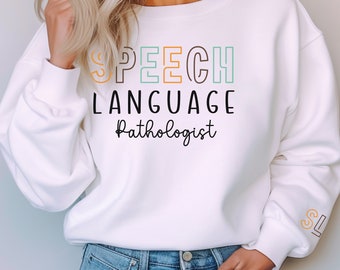 Puede incluir: Sudadera blanca con el texto "SPEECH LANGUAGE Pathologist" en letras de colores con contorno. Las letras "SL" están bordadas en la manga.