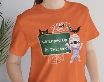May include: Orange t-shirt featuring a Halloween graphic. The design includes a black cat, a mummy, a bat, and a chalkboard that reads "Wrapped Up in Teaching." A white spider web is also featured in the design. The words "Happy Halloween" are written above the chalkboard.