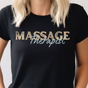 Op de afbeelding: Zwart T-shirt met de tekst "Massage Therapist" in een verweerd, vintage lettertype. Het woord "Massage" is in lichtbruin, en het woord "Therapist" is in lichtblauw.