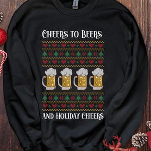 Op de afbeelding: Zwart sweatshirt met een feestelijk ontwerp. Het ontwerp bevat de tekst "CHEERS TO BEERS AND HOLIDAY CHEERS" boven en onder een patroon van harten, kerstbomen en bierpullen. De bierpullen zijn gevuld met goudkleurig bier en bedekt met wit schuim.