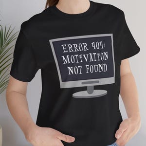 Op de afbeelding: Zwart T-shirt met een computerbeeldschermafbeelding met de tekst "ERROR 404: MOTIVATION NOT FOUND" in het wit. Het scherm heeft een grijze rand en standaard. Het shirt heeft korte mouwen.