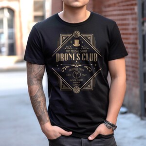 May include: A black t-shirt featuring a gold-colored graphic design with the text THE FEUDAL SPIRIT DRONES CLUB, MEMBERS ONLY, ESTD 1920, DOVER STREET, MAYFAIR, LONDON, and an illustration of a top hat and a martini glass within a geometric frame.