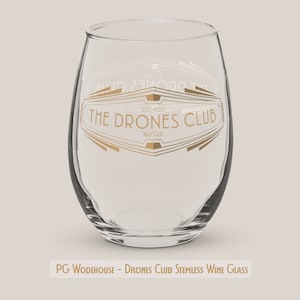 May include: A clear stemless wine glass with a gold graphic that reads "The Drones Club, Est. 1920, Mayfair".