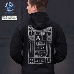 Puede incluir: Sudadera con capucha negra con un gr&aacute;fico blanco en la espalda. El dise&ntilde;o incluye el texto "LIBER AL VEL LEGIS" en una fuente estilizada. La sudadera tiene un cuello alto y solo una impresi&oacute;n en la espalda.