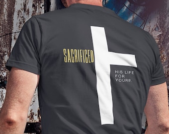 May include: Dark gray t-shirt with a large white cross graphic on the back. The shirt features the text "SACRIFICED" in gold and "HIS LIFE FOR YOURS" in white. The model is wearing blue jeans.