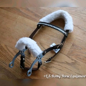 May include: A brown leather horse halter with a white fleece noseband and browband. The halter has a row of rhinestones on the browband and a silver bit with a white fleece cheek piece.