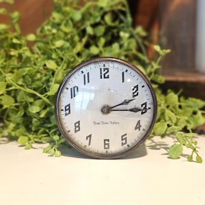 May include: An antique clock with a white face, black numerals, and black hands. The clock has a metal casing and the words "True Time Tellers" are printed on the face. The clock is set to approximately 3:15.