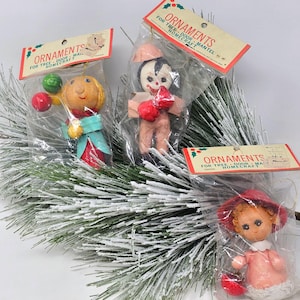 May include: Three vintage Christmas ornaments in plastic bags. One ornament is a clown with red gloves and a hat. Another is a girl in a pink dress and red hat. The third is a doll holding colorful balls. Each has a tag that reads "ORNAMENTS FOR TREE DOOR MANTEL HOMECRAFT".
