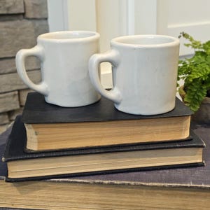 May include: Two off-white ceramic mugs with rounded handles sit atop a stack of vintage books. The mugs have a simple, classic design. The books have dark blue covers and aged, yellowed pages, suggesting a well-loved collection.