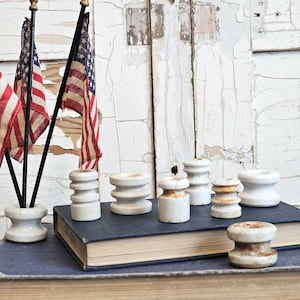 May include: A collection of white ceramic electrical insulators with ribbed sides, some showing brown staining. Two small American flags are inserted into one of the insulators. The items are arranged on top of a dark blue hardcover book.