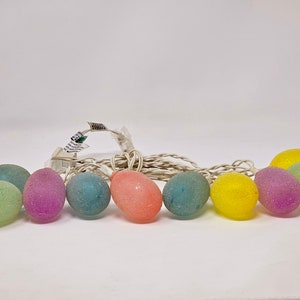 May include: A string of Easter egg lights in various colors, including green, purple, blue, yellow, and pink. The egg-shaped lights are strung on a white cord and are illuminated. The lights are designed for Easter decorations.