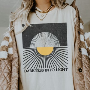 May include: A white t-shirt with a black and white graphic of a sun with the text "Darkness into Light".