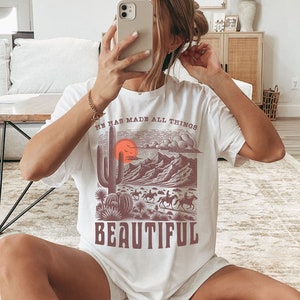K&ouml;nnte beinhalten: Wei&szlig;es T-Shirt mit einem grafischen Druck einer W&uuml;stenlandschaft mit einem Kaktus, Bergen und einer untergehenden Sonne. Der Text "He has made all things beautiful" ist auf das Shirt gedruckt.