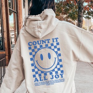 May include: A beige hoodie with a blue checkered pattern and a blue smiley face graphic. The text "COUNT IT ALL JOY" is printed below the graphic. The hoodie is being worn by a person.