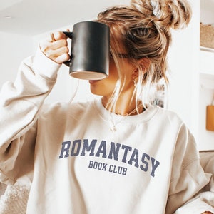 May include: A light beige sweatshirt with the text "Romantasy Book Club" in blue lettering.