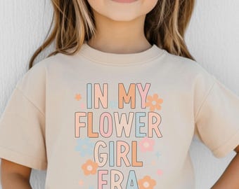 May include: A young person wearing heart-shaped sunglasses and a beige t-shirt. The t-shirt has the text "IN MY FLOWER GIRL ERA" in pastel colors. The word "NATURAL" is printed in a white rectangle at the bottom right.