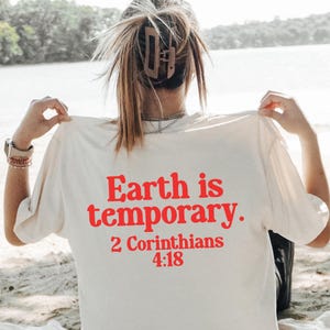 May include: Ivory t-shirt with red text that reads 'Earth is temporary. 2 Corinthians 4:18.' The person is sitting on a white blanket with food and drinks, including watermelon, tomatoes, and donuts. The background includes a lake and trees.