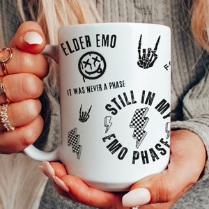 May include: White ceramic mug with the text "ELDER EMO" and "IT WAS NEVER A PHASE" in black. The mug features a smiley face with an X through the eyes, a skeleton hand, and a checkered lightning bolt design. The words "STILL IN MY EMO PHASE" are also printed on the mug.