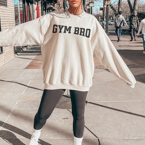May include: Sand-colored sweatshirt with the text "GYM BRO" in black block letters. The sweatshirt is paired with black leggings and white sneakers. The image is taken outdoors on a sunny day.