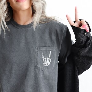 May include: A dark gray long-sleeved t-shirt with a pocket on the left side. The pocket has a white skeleton hand making a rock and roll sign.