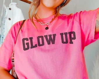 Puede incluir: Una camiseta rosa brillante con las palabras "GLOW UP" impresas en letras oscuras grises. La camiseta es de un color vibrante y parece estar hecha de material suave. La palabra "CRUNCHBERRY" está en un óvalo blanco en la parte inferior de la imagen.