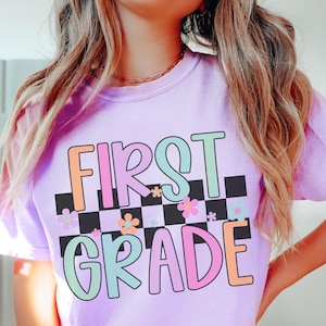 May include: A light purple t-shirt with the words "FIRST GRADE" in a colorful, retro font. The text is set against a black and white checkered background with flower accents. The shirt is a comfortable, casual style.