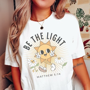 May include: White t-shirt with a yellow sun graphic and the text "Be the Light" with the Bible verse "Matthew 5:14" underneath.