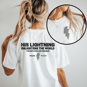 May include: White t-shirt with the text "HIS LIGHTNING ENLIGHTENS THE WORLD" and "PSALMS 97:4 KJV" printed in black. The shirt features a checkered lightning bolt graphic. The shirt is a casual wear item.