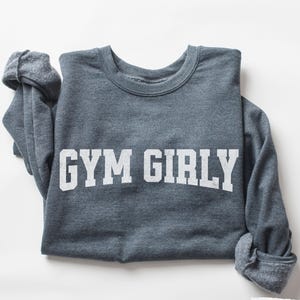 May include: Dark grey heather sweatshirt with the text "GYM GIRLY" in white block letters. The long-sleeved crew neck top is folded, with the sleeves rolled up. The words "DARK GREY HEATHER" are printed in a small white font at the bottom right.