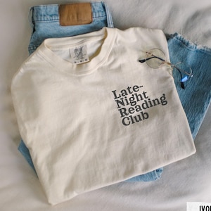 Puede incluir: Una camiseta blanca con el texto "Late-Night Reading Club" impreso en negro. La camiseta est&aacute; doblada y colocada sobre un par de pantalones vaqueros azules.