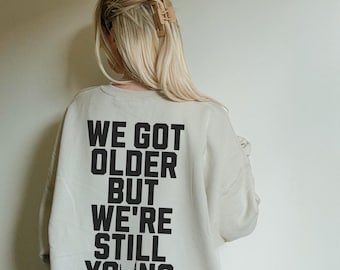 May include: A white sweatshirt with black text that reads "We got older but we're still young" with a rock and roll hand gesture.