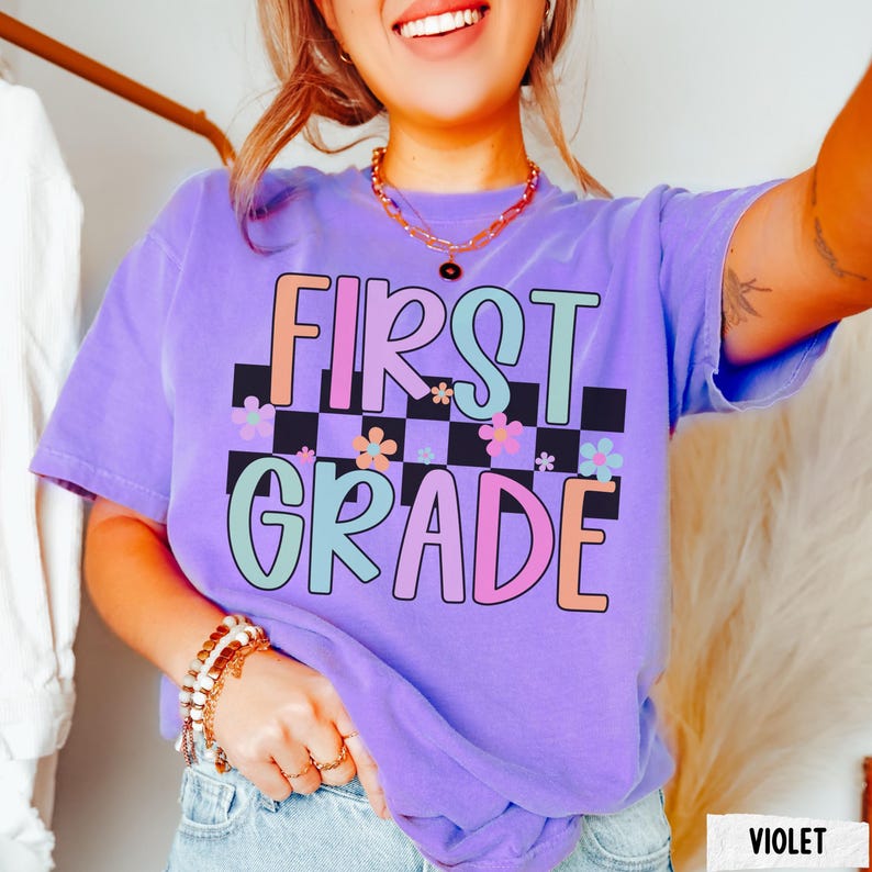 K&ouml;nnte beinhalten: Violettes T-Shirt mit dem Schriftzug "FIRST GRADE" in bunten Buchstaben. Der Text ist vor einem schwarz-wei&szlig; karierten Hintergrund mit floralen Akzenten platziert. Das Shirt ist ein l&auml;ssiger, bequemer Stil.