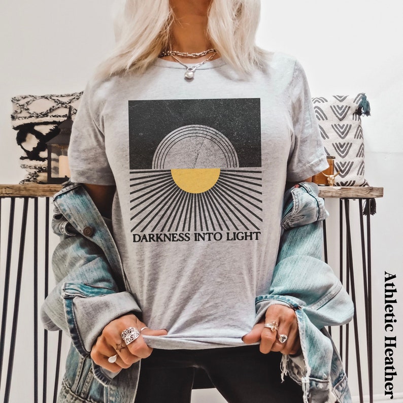 May include: A gray t-shirt with a graphic design of a yellow sun rising over a black background. The text "DARKNESS INTO LIGHT" is printed below the sun.
