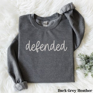 May include: Dark grey heather sweatshirt with the word "defended" in a cursive font across the front. The long sleeves are cuffed. The sweatshirt is made of a soft material. The tag reads "Gildan".