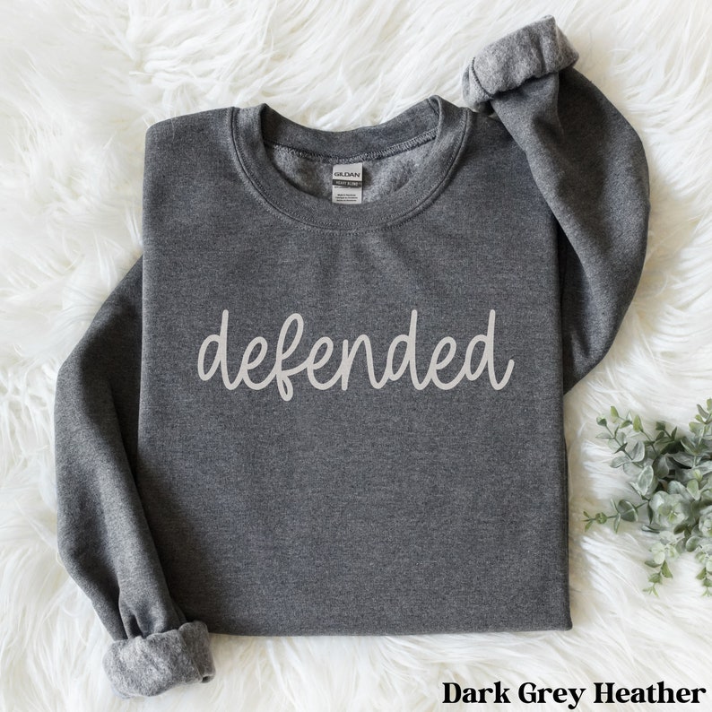 May include: Dark grey heather sweatshirt with the word "defended" in a cursive font across the front. The long sleeves are cuffed. The sweatshirt is made of a soft material. The tag reads "Gildan".