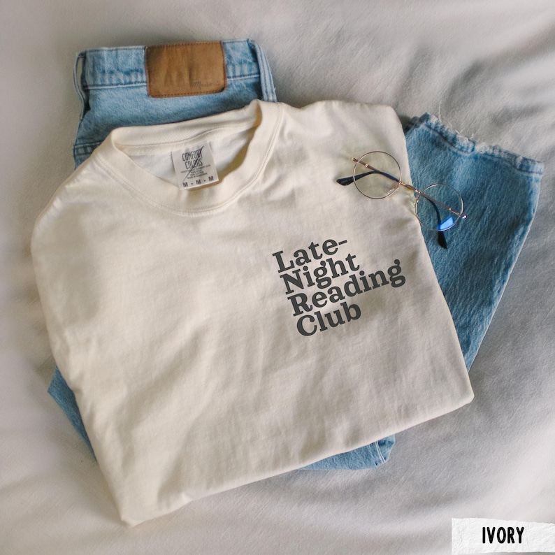 Puede incluir: Una camiseta blanca con el texto "Late-Night Reading Club" impreso en negro. La camiseta est&aacute; doblada y colocada sobre un par de pantalones vaqueros azules.