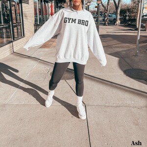 May include: Light grey sweatshirt with the text "GYM BRO" in black block letters. The sweatshirt is oversized and worn with black leggings and white sneakers. The image is taken outdoors on a sunny day.