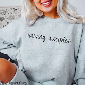 May include: A light gray sweatshirt with the words "raising disciples" in black script. The sweatshirt is a sport grey color and has a crew neck.