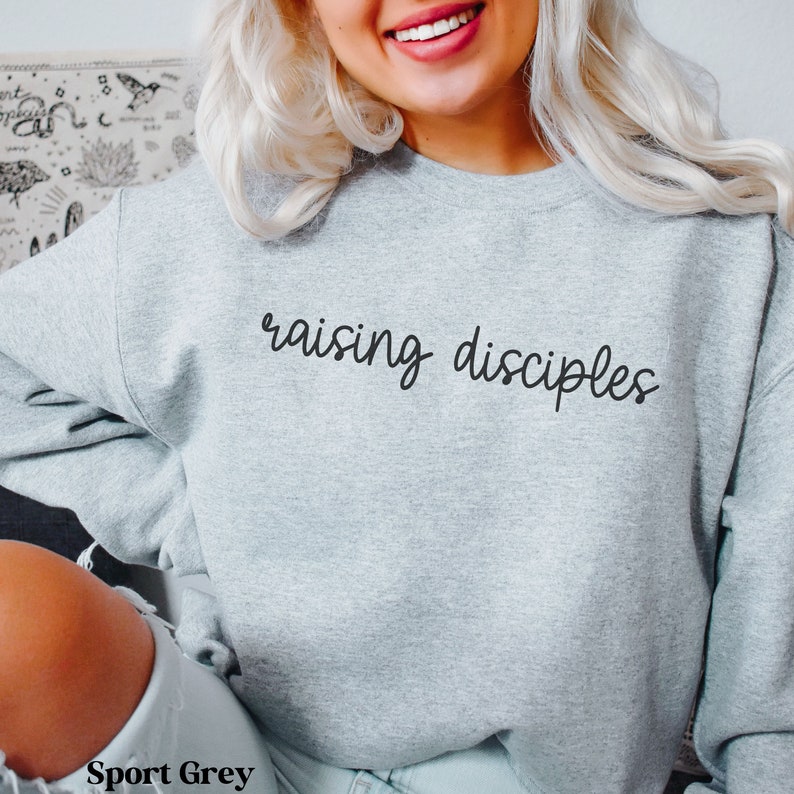 May include: A light gray sweatshirt with the words "raising disciples" in black script. The sweatshirt is a sport grey color and has a crew neck.