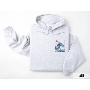 May include: Light gray hoodie with a drawstring hood. The front features a rectangular graphic of a wave and a red sun. The word "ASH" is printed in a white rectangle at the bottom right.
