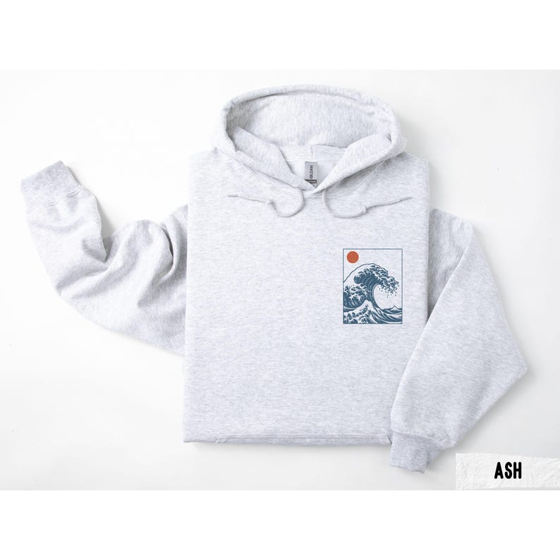 May include: Light gray hoodie with a drawstring hood. The front features a rectangular graphic of a wave and a red sun. The word "ASH" is printed in a white rectangle at the bottom right.