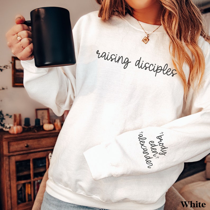 May include: White sweatshirt with the words "raising disciples" in black script across the chest. The sleeve has the names "Brody, Eden, Alexander" in black script. A black mug is held in the hand.